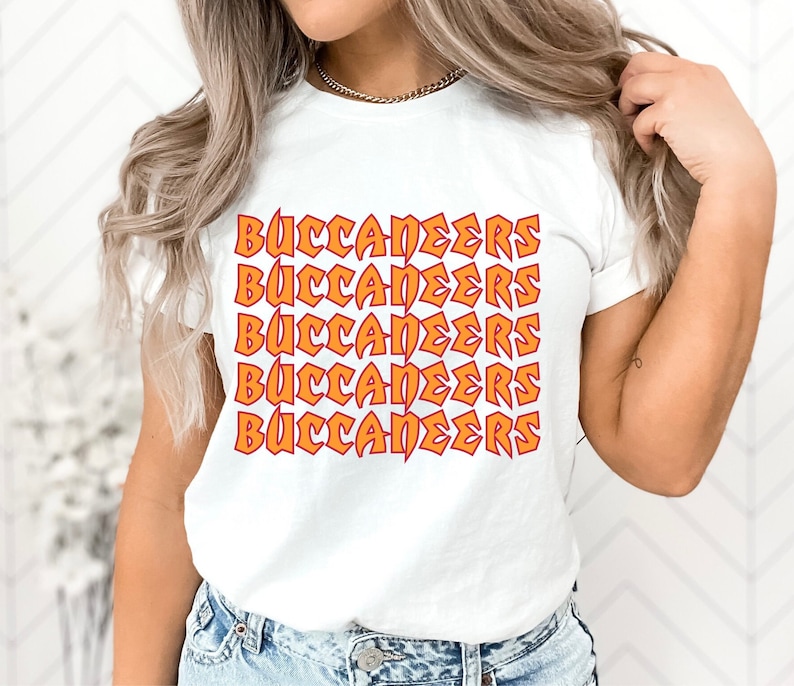 May include: White t-shirt with the word "BUCCANEERS" in orange and red repeating across the front. The text has a bold, slightly distressed design. The shirt has short sleeves and a crew neck.