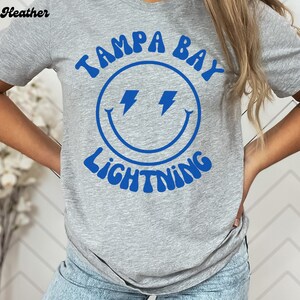 May include: Gray t-shirt with a blue smiley face graphic and the text "Tampa Bay Lightning" in a retro font.