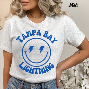 May include: A gray t-shirt with a blue smiley face graphic and the text "Tampa Bay Lightning" in a retro font.