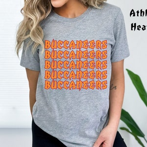 May include: A gray t-shirt with the word "BUCCANEERS" in orange and red lettering repeated five times. The shirt is labeled "Athletic Heather" in black text.
