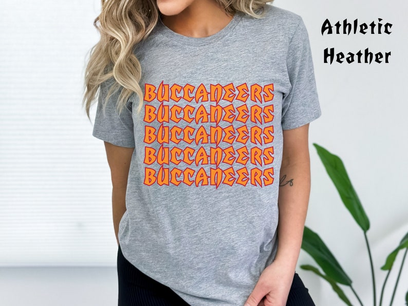 May include: A gray t-shirt with the word "BUCCANEERS" in orange and red lettering repeated five times. The shirt is labeled "Athletic Heather" in black text.