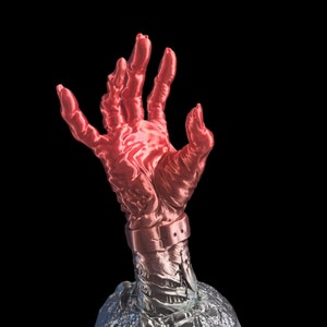 May include: A red and silver zombie hand sculpture with a metal wristband. The hand is raised in a grasping motion and is set on a textured base.