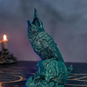 May include: A teal green 3D printed bird sculpture perched on a skull. The bird has its beak open and its wings are spread out. The sculpture is on a round base.