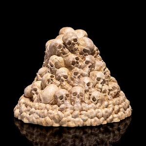 Puede incluir: Una pila de pequeños cráneos de cerámica blanca dispuestos en forma de cono. Los cráneos están todos orientados en diferentes direcciones y están apilados unos sobre otros. La base de la pila está rodeada por un anillo de cráneos de cerámica blanca más pequeños.