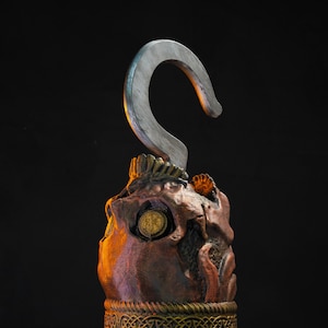 May include: A decorative pirate hook with a skull and crossbones design. The hook is made of metal and has a wooden handle. The skull is painted in shades of brown and red, and the crossbones are painted in gold. The hook is mounted on a wooden base with a gold trim.
