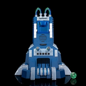 May include: A blue and silver 3D printed robot with green tubes on top. The robot has a face with black eyes and a mouth. It is standing on a black surface with a green die in front of it.