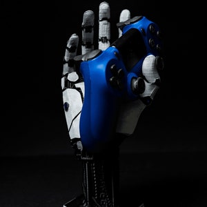 May include: A 3D printed robotic hand holding a blue video game controller. The hand is white and gray, and the controller has black buttons.
