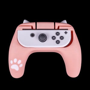 May include: A pink and white Nintendo Switch controller with paw print grips. The controller has a white faceplate with black buttons and a pink back.