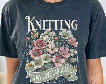 May include: Dark gray t-shirt with the text "Knitting is my love language" above a floral design in pink, white, and green. The shirt has short sleeves and a relaxed fit, perfect for casual wear.