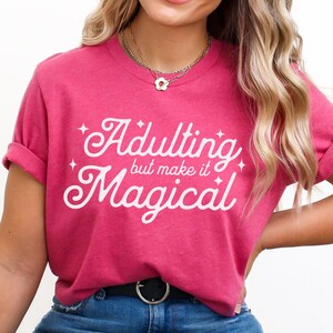 May include: Heather raspberry t-shirt with rolled sleeves and the text "Adulting but make it Magical" in white script. The model is wearing blue jeans with ripped knees and a black belt. A small feather graphic is in the upper right corner.