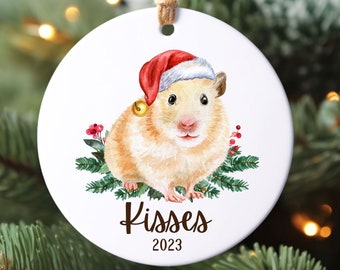 May include: A white ceramic Christmas ornament featuring a watercolor illustration of a hamster wearing a Santa hat. The hamster is surrounded by green pine branches and red berries. The ornament has the text "Kisses 2023" printed on it.