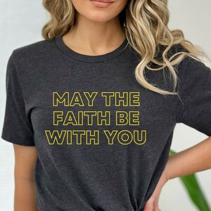 May include: A dark grey heather t-shirt with the text "MAY THE FAITH BE WITH YOU" in yellow.