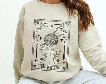 May include: A beige sweatshirt with a black graphic of a yarn ball and knitting needles. The graphic is in the style of a tarot card and says "The Knitter".