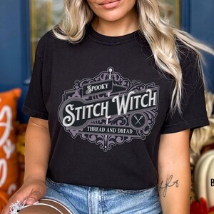 May include: A black t-shirt with a graphic design of a vintage logo that says "Spooky Stitch Witch Thread and Dread". The logo is in purple and gray with a floral design. The model is wearing a beige beanie and blue jeans.