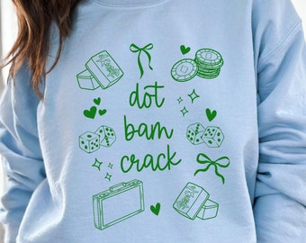 Może przedstawiać: Jasno błękitna bluza z zielonym nadrukiem graficznym z tekstem "dot bam crack" i różnymi obrazkami, w tym kostkami do gry, kartami do gry i walizką.