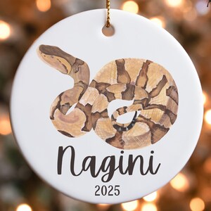 May include: White ceramic ornament featuring a coiled snake illustration in shades of brown and tan. The name "Nagini" is written in black script below the snake, with "2025" beneath it. A gold string is attached for hanging.