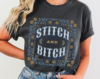 May include: A dark gray t-shirt with a floral design and the text "Stitch and Bitch" in blue and yellow lettering.