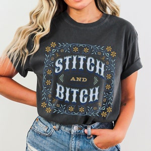 May include: A dark gray t-shirt with a floral design and the text "Stitch and Bitch" in blue and yellow lettering.