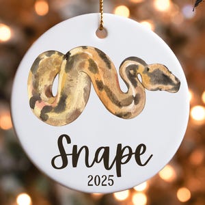 May include: White ceramic ornament featuring a watercolor illustration of a snake in shades of brown and tan. The word "Snape" is written in black script below the snake, with "2025" beneath it. A gold string is attached for hanging.
