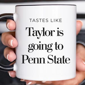 May include: White ceramic mug with a black handle and rim. The mug features the text "TASTES LIKE Taylor is going to Penn State" in a bold, black font. The mug is held by a person.