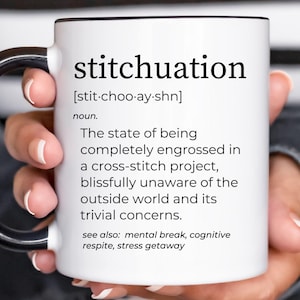 May include: White ceramic mug with a black handle and rim, featuring the word "stitchuation" and its definition. The definition describes being engrossed in a cross-stitch project. The mug is held by a person.