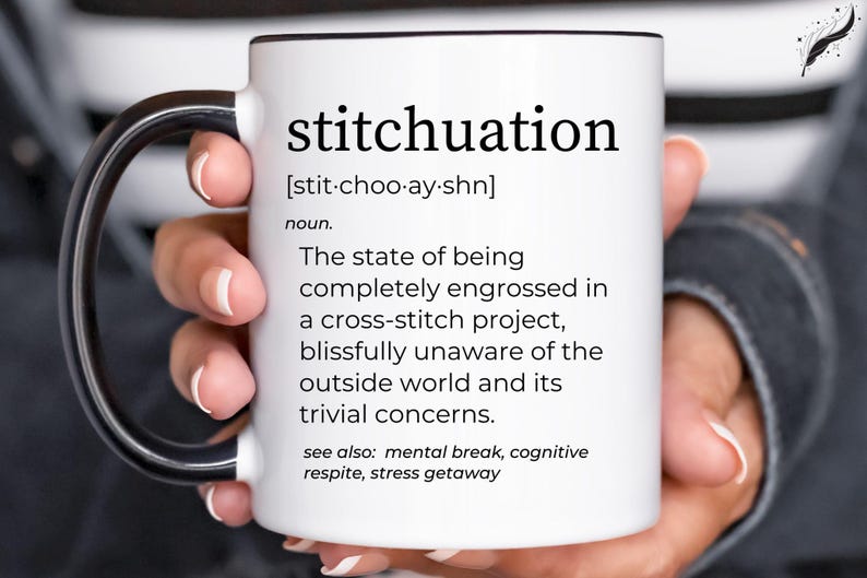 May include: White ceramic mug with a black handle and rim, featuring the word "stitchuation" and its definition. The definition describes being engrossed in a cross-stitch project. The mug is held by a person.