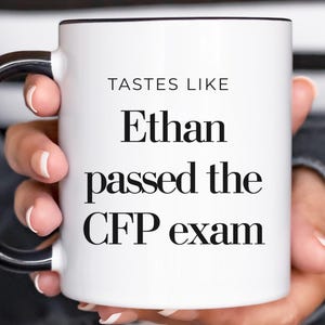 May include: White ceramic mug with a black handle and rim. The mug features the text "TASTES LIKE" above "Ethan passed the CFP exam" in a bold, black font. The mug is held by a person.