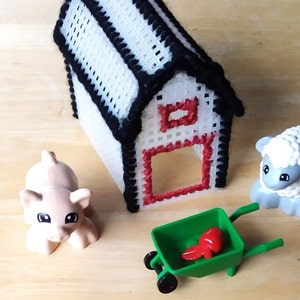 May include: A miniature farm scene featuring a white and black barn with red accents, a pink pig, a sheep, and a green wheelbarrow with a red object inside. The toys are arranged on a light brown wooden surface.