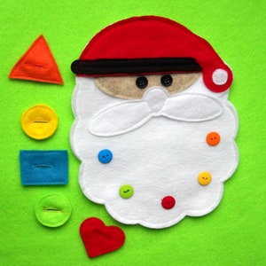 May include: A felt Santa Claus face with a red hat and white beard, adorned with colorful buttons. Various felt shapes, including a triangle, circle, rectangle, and heart, are scattered around the Santa face on a green background. The shapes are orange, yellow, blue, green, and red.