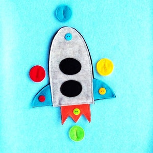 May include: A light blue felt background features a gray rocket ship with black portholes and orange flames. Colorful buttons in red, yellow, green, and blue are scattered around the rocket, creating a playful, space-themed design.