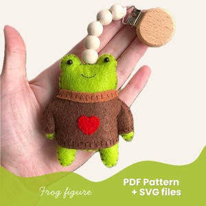 May include: A handmade green felt frog figure wearing a brown sweater with a red heart. The frog is attached to a wooden pacifier clip with wooden beads. The image also includes the text "Frog figure" and "PDF Pattern + SVG files".