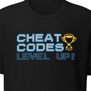 Może przedstawiać: Czarny T-shirt z niebiesko-białym pikselowanym nadrukiem z napisem "CHEAT CODES LEVEL UP!" i żółtym pikselowanym trofeum.