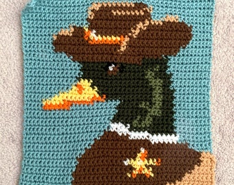 May include: Crocheted square featuring a duck wearing a brown cowboy hat with orange accents. The duck is wearing a brown vest with a yellow star and a white collar. The background is a light blue color.