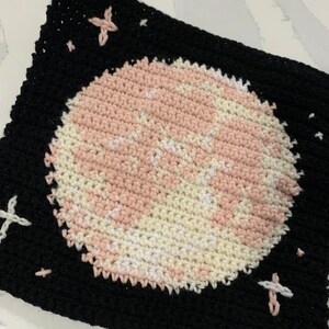 Puede incluir: Un cuadrado tejido a crochet con un diseño de planeta rosa y blanco sobre un fondo negro. El planeta está rodeado de pequeñas estrellas blancas.