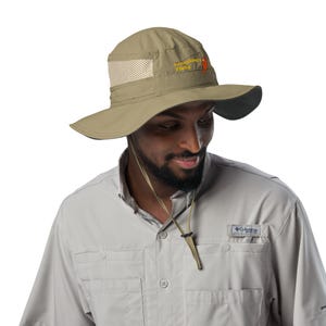 May include: A khaki-colored sun hat with a wide brim and mesh ventilation. The hat features the text "Something's Fishy" in yellow and an orange fish graphic. It has an adjustable chin strap.