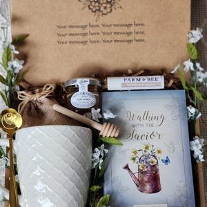 May include: A gift box with a white mug, honey, a book titled "Walking with the Savior," and a vanilla bean lip balm. The mug has a textured pattern, and the book features a floral illustration. The box is decorated with white flowers.