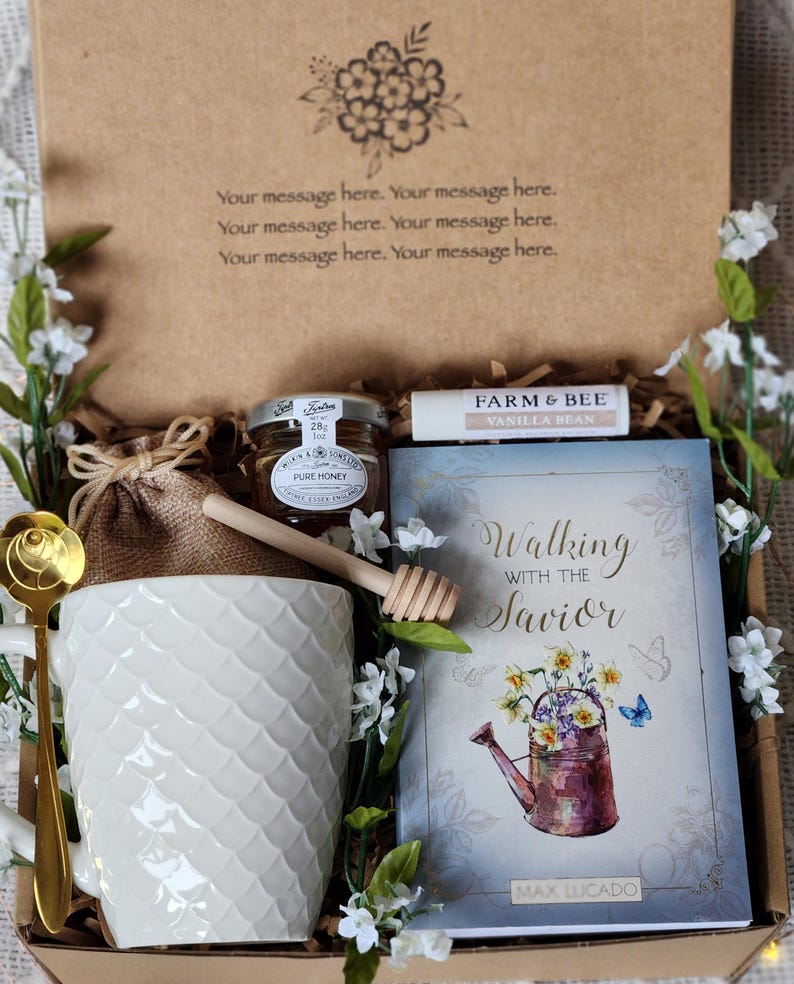 May include: A gift box with a white mug, honey, a book titled "Walking with the Savior," and a vanilla bean lip balm. The mug has a textured pattern, and the book features a floral illustration. The box is decorated with white flowers.