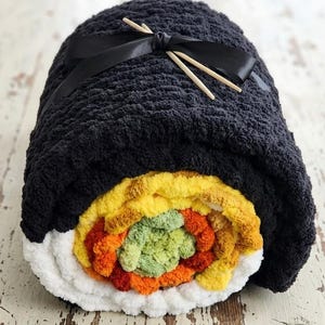 May include: A plush, black blanket rolled to resemble sushi. The blanket features a colorful, textured filling with shades of yellow, orange, and green. A black ribbon and two wooden chopsticks are tied on top, completing the sushi roll design.