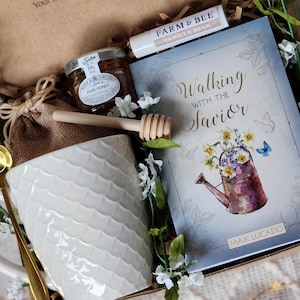 May include: A gift box containing a white ceramic mug, a jar of honey, a wooden honey dipper, a lip balm, a book titled "Walking with the Savior", and a gold spoon. The mug has a textured scale pattern.