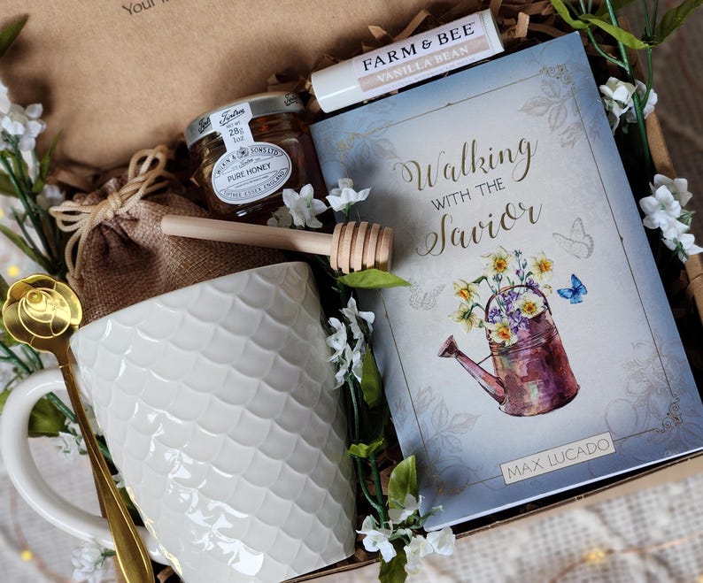 May include: A gift box containing a white ceramic mug, a jar of honey, a wooden honey dipper, a lip balm, a book titled "Walking with the Savior", and a gold spoon. The mug has a textured scale pattern.