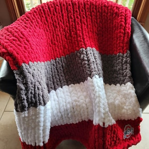 May include: A chunky knit blanket in red, gray, white, and brown stripes, draped over a chair. The blanket features a team logo in the corner. The thick yarn creates a textured, cozy appearance, perfect for snuggling.