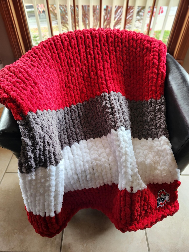 May include: A chunky knit blanket in red, gray, white, and brown stripes, draped over a chair. The blanket features a team logo in the corner. The thick yarn creates a textured, cozy appearance, perfect for snuggling.