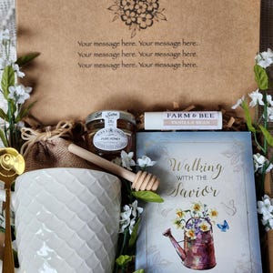 May include: A gift box with a white textured mug, honey, lip balm, and a book titled "Walking with the Savior." The box has a floral design and the text "Your message here." Decorative flowers surround the items.