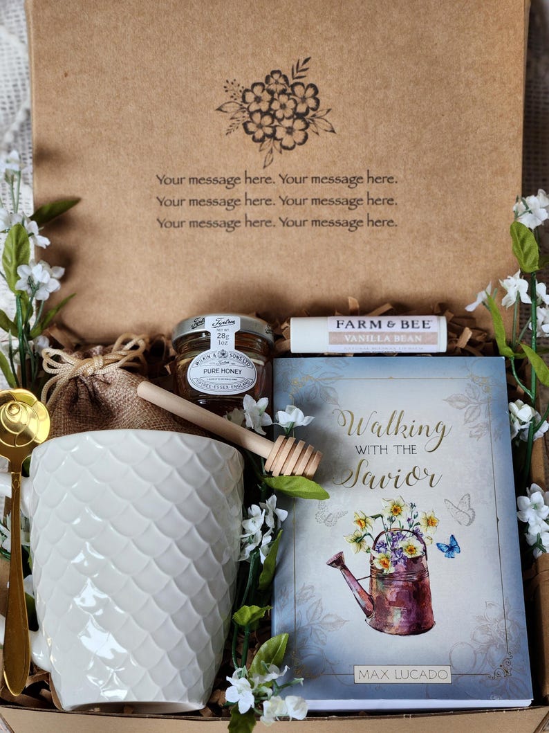 May include: A gift box with a white textured mug, honey, lip balm, and a book titled "Walking with the Savior." The box has a floral design and the text "Your message here." Decorative flowers surround the items.