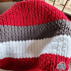 May include: A handmade, chunky knit blanket in red, gray, white, and brown stripes. The blanket features an Ohio State logo patch in the corner. The soft, plush blanket is perfect for snuggling up on the couch or bed.