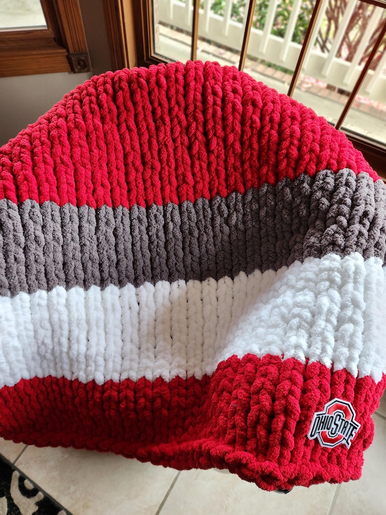 May include: A handmade, chunky knit blanket in red, gray, white, and brown stripes. The blanket features an Ohio State logo patch in the corner. The soft, plush blanket is perfect for snuggling up on the couch or bed.