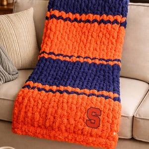 May include: A chunky knit blanket in orange and navy blue, featuring horizontal stripes and the letter "S" in a patch. The blanket is draped over a beige sofa, suggesting a cozy home decor item.