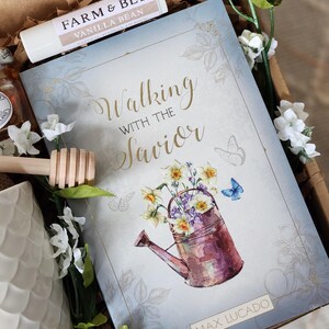 May include: A gift box contains a book titled "Walking with the Savior" by Max Lucado, a lip balm, and a honey dipper. The book cover features a watercolor illustration of a watering can filled with flowers. White flowers and greenery surround the items.