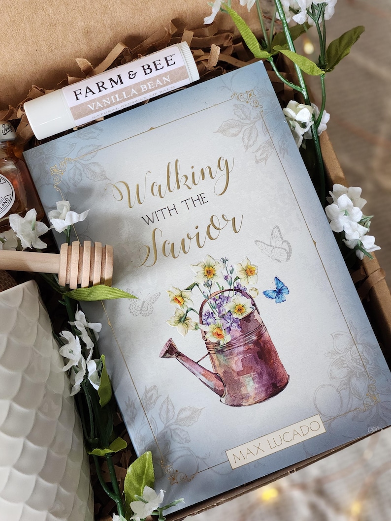 May include: A gift box contains a book titled "Walking with the Savior" by Max Lucado, a lip balm, and a honey dipper. The book cover features a watercolor illustration of a watering can filled with flowers. White flowers and greenery surround the items.
