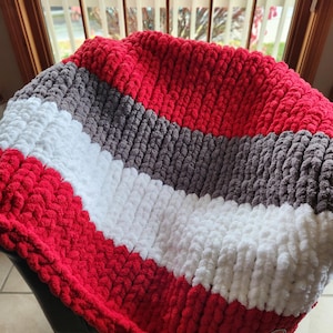 May include: A chunky knit blanket in red, white, and gray stripes. The blanket is made of a soft, textured material and is folded over a dark surface. The blanket appears to be handmade and is suitable for home decor.